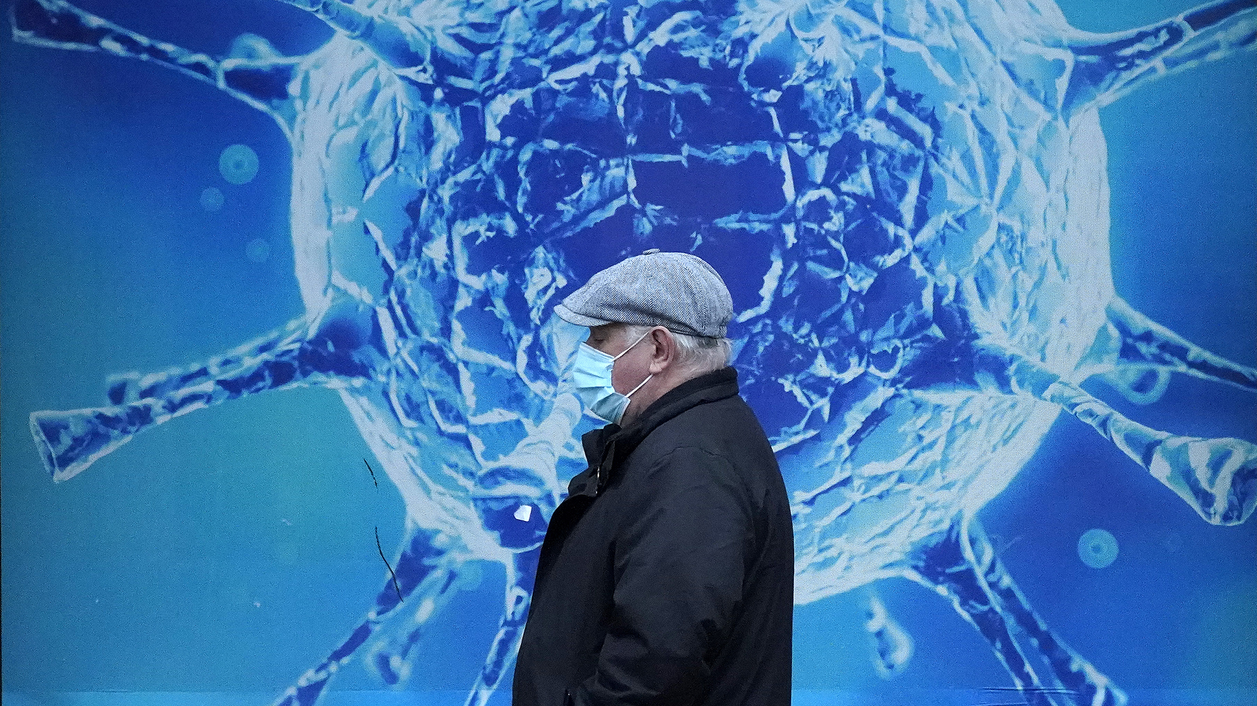 A man wearing a face mask walks past a poster of a coronavirus, symbolizing the ongoing impact of the pandemic.